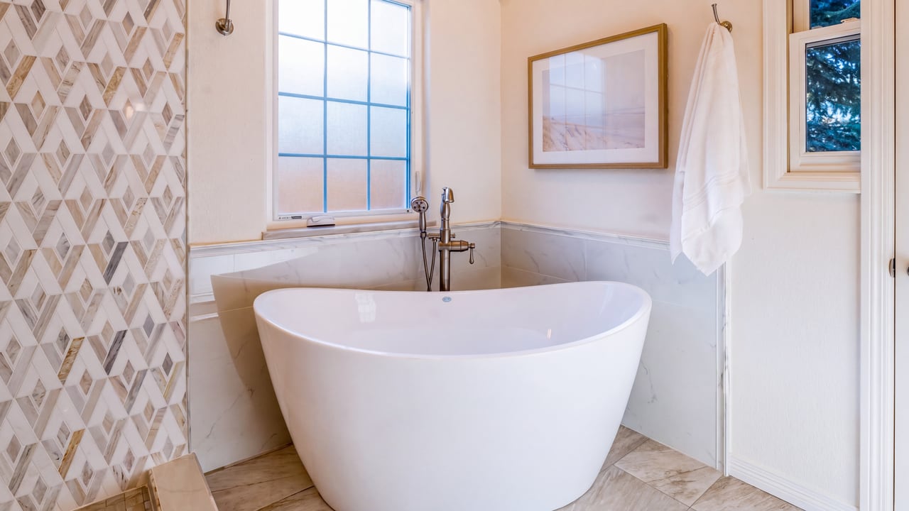 Freestanding white bathtub beside a window and patterned tile wall